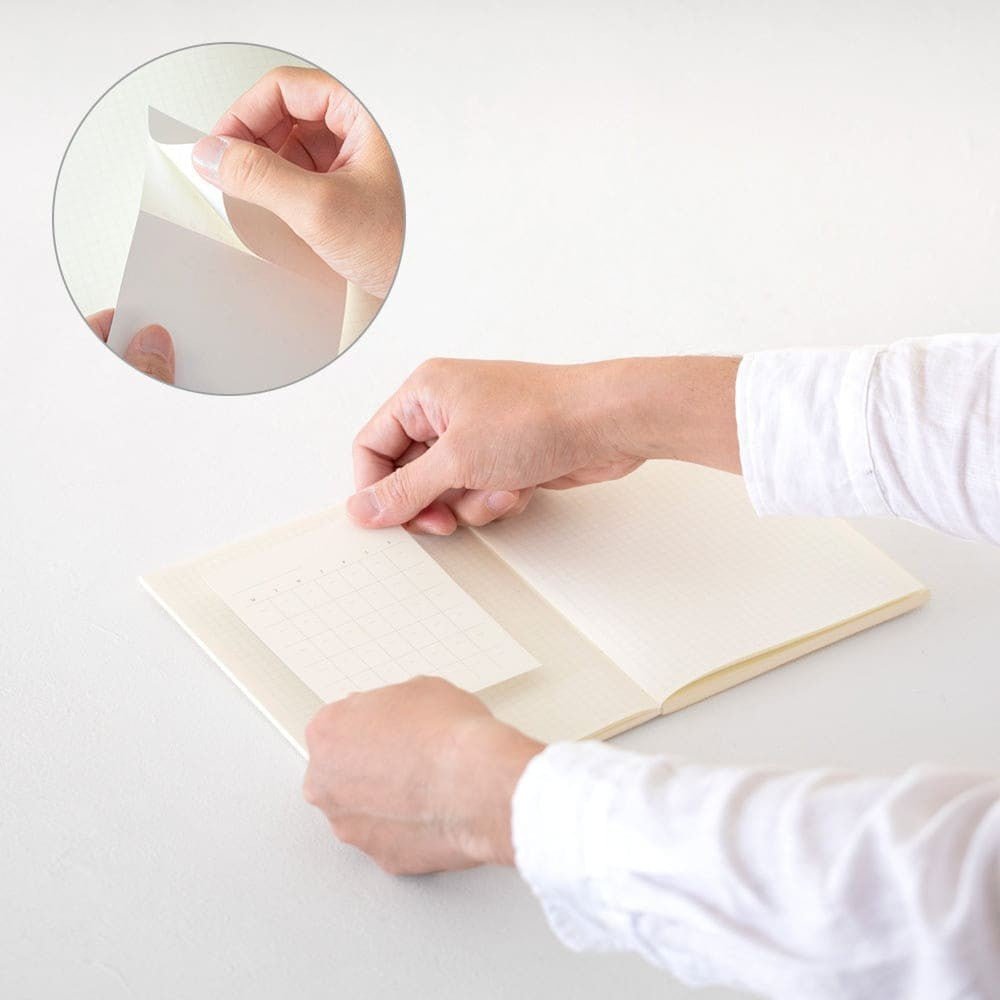 MD Diary Sticker Free från Midori - Händer sätter ett klistermärkesark med ett månadsuppslag i en anteckningsbok. Tidformera