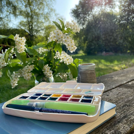 En låda med akvarellfärger ligger ute på ett bord. Vacker natur runt. - Tidformera