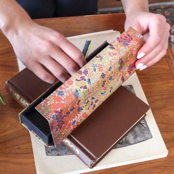 Paperblanks pennskrin. Blommigt pennfack öppnas av händer. Pennfack ligger på ett läsebok.  - Tidformera