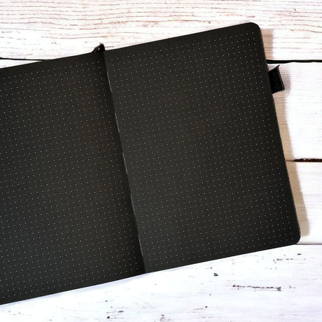 En öppen skrivbok ur serien Black out Journal från Paper24. Boken har svarta blad som är dotted. - Tidformera