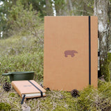 Anteckningsbok Wildlife Dotted A4 Brown Bear från Dingbats*. Två anteckningsböcker, varav en är A4 och den andra är A6 storlek, med det bruna björnmotivet. De finns i skogen bland mossa och kottar. Bakom den lilla boken finns en kåsa. - Tidformera