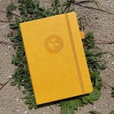 Dotted Notebook A5 160 GSM Smiling sun från Paper24. Gul anteckningsbok med motiv med en leende sol. Skrivboken ligger ute i solen på växter. - Tidformera