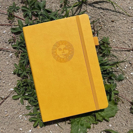 Dotted Notebook A5 160 GSM Smiling sun från Paper24. Gul anteckningsbok med motiv med en leende sol. Skrivboken ligger ute i solen på växter. - Tidformera